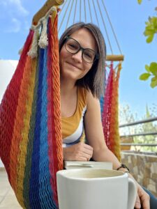 Vic_Profile Victoria with a slight smile looking at the camera, sitting outside in a multi-colored hammock chair. Background is a blue sky with some plants in te distance, and Victoria wear green glasses, a yellow and white tank top, and holds a book on her lap, with a coffee mug just in front.