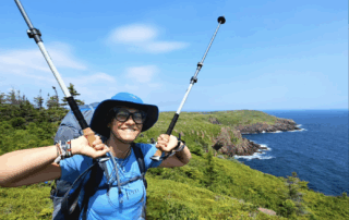 Victoria standing with walking sticks raised in her hands on teh side of the East Coast Trrail in Newfoundland, on a green trail and the ocean and moutains pictured in the background. She is smiling and wearing a blue hat, light blue tshirt and a heavy backpack on her back.